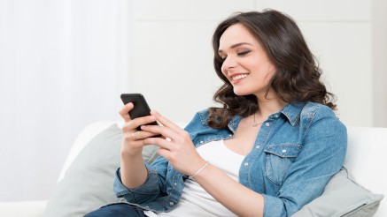 GesundheitsApp: Junge Frau sitzt mit dem Smartphone auf dem Sofa und lädt sich die App herunter.